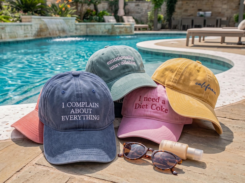 May include: A collection of five baseball caps in blue, green, pink, yellow, and coral. The blue cap reads I COMPLAIN ABOUT EVERYTHING, the green cap reads emotionally unavailable, the pink cap reads I need a Diet Coke, and the yellow cap reads california.