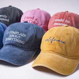 May include: A collection of six baseball caps in various colors including black, pink, red, blue, mustard yellow, and teal. Each cap features embroidered text on the front, such as Heavily Caffeinated, I need a Diet Coke, I could be meaner, and I COMPLAIN ABOUT EVERYTHING.