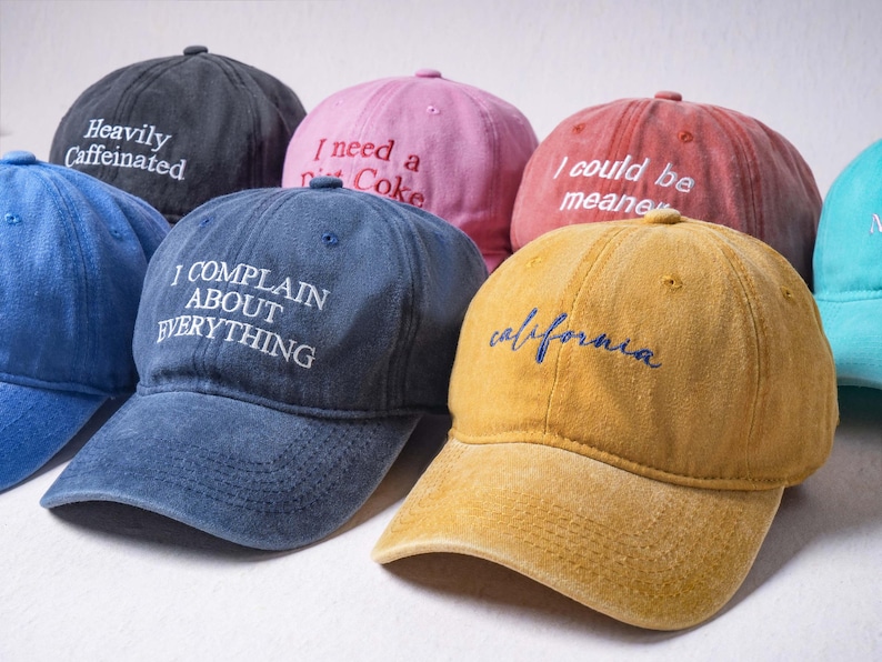 May include: A collection of six baseball caps in various colors including black, pink, red, blue, mustard yellow, and teal. Each cap features embroidered text on the front, such as Heavily Caffeinated, I need a Diet Coke, I could be meaner, and I COMPLAIN ABOUT EVERYTHING.