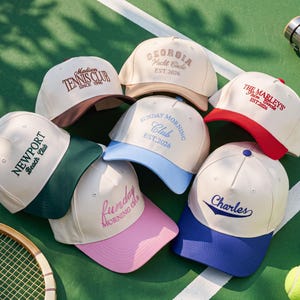 Puede incluir: Una colección de siete gorras de béisbol con coronas de color blanco roto y viseras coloridas. Cada gorra presenta un texto bordado único que incluye Madison Tennis Club, Georgia Yacht Circle, The Marley's Palm Circle, Sunday Morning Club, Funday Morning Club y Charles.