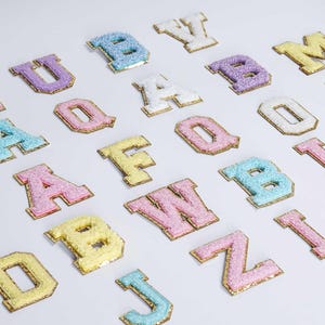 以下が含まれることがあります： ピンク、ブルー、イエロー、パープルなどのパステルカラーの刺繍入りバーシティレターパッチの詰め合わせ。 各文字は金色の縁取りとふわふわした質感で、カスタマイズプロジェクトに最適です。