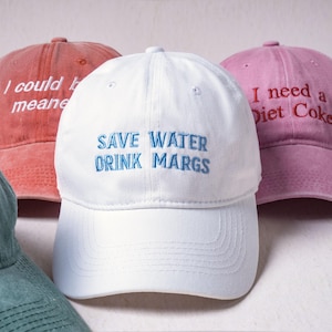 May include: A collection of four baseball caps in white, pink, red, and green. The white cap features blue embroidered text reading SAVE WATER DRINK MARGS. The pink cap has red text reading I need a Diet Coke. The red cap has white text.
