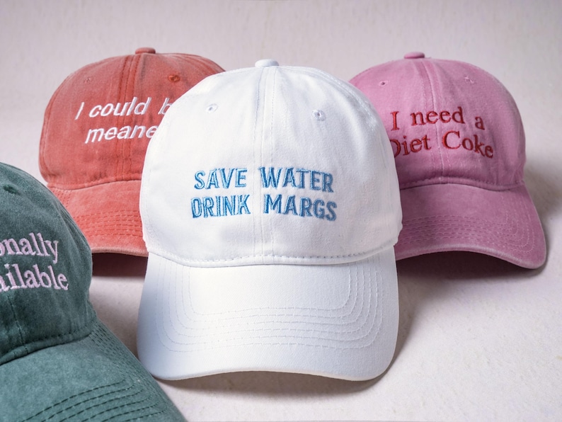 May include: A collection of four baseball caps in white, pink, red, and green. The white cap features blue embroidered text reading SAVE WATER DRINK MARGS. The pink cap has red text reading I need a Diet Coke. The red cap has white text.