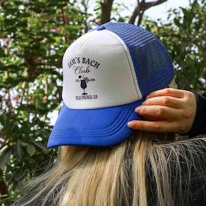 May include: A blue and white trucker hat with a black logo that says "Sam's Bach Club est. 2023 Palm Springs, CA".