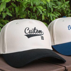 May include: Two baseball caps with a cream-colored crown and a black brim. One cap has the word "Custom" embroidered in black with the number "18" below. The other cap has the words "Embroider Here" embroidered in blue.
