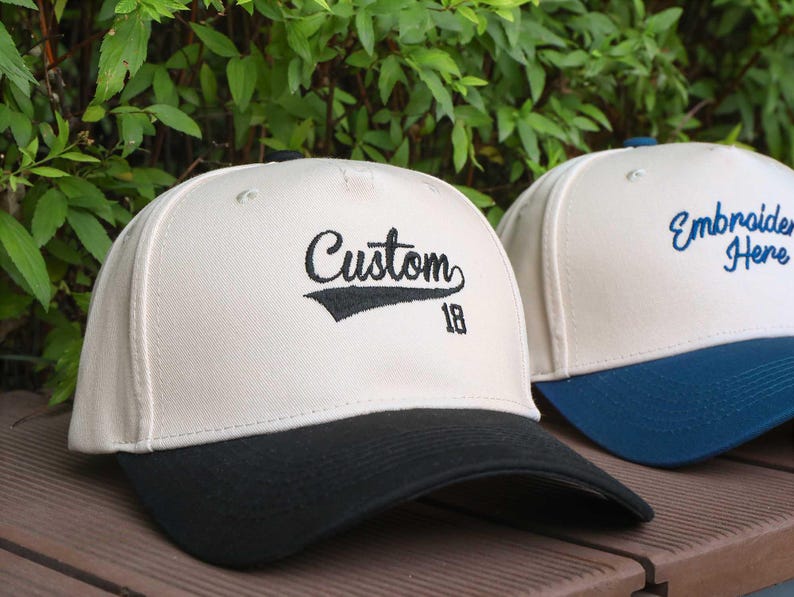 May include: Two baseball caps with a cream-colored crown and a black brim. One cap has the word "Custom" embroidered in black with the number "18" below. The other cap has the words "Embroider Here" embroidered in blue.