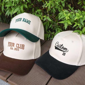 May include: Three baseball caps with custom embroidery. One cap has "YOUR NAME" in green, another says "YOUR CLUB Est. 2025" in brown, and the third says "Custom 18" in black. Each cap has a different colored brim.
