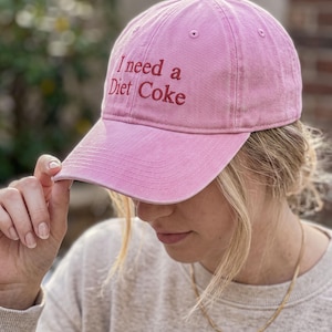 May include: A pink baseball cap with a curved brim and an embroidered red text on the front that reads I need a Diet Coke. The cap features a button on the top center and stitched eyelets for ventilation, showcasing a classic six-panel construction.
