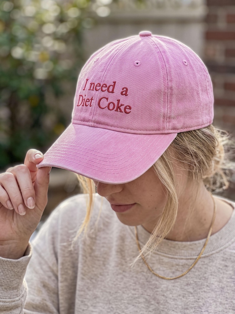 May include: A pink baseball cap with a curved brim and an embroidered red text on the front that reads I need a Diet Coke. The cap features a button on the top center and stitched eyelets for ventilation, showcasing a classic six-panel construction.