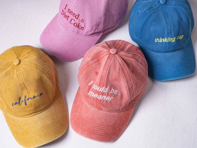 May include: Four baseball caps in mustard yellow, light pink, coral red, and blue. The yellow cap features the word "california" in blue. The pink cap says "I need a Diet Coke" in red. The coral cap says "I could be meaner" in white. The blue cap says "thinking cap" in yellow.