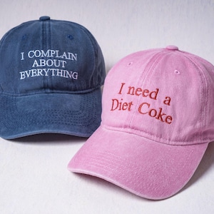May include: A navy blue baseball cap with white embroidered text reading I COMPLAIN ABOUT EVERYTHING and a pink baseball cap with red embroidered text reading I need a Diet Coke. Both caps feature a curved brim and a six-panel construction.