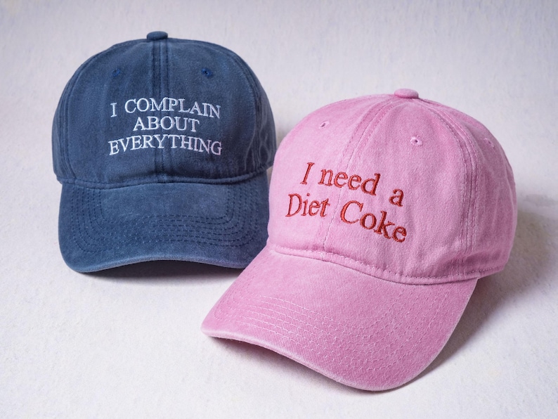 May include: A navy blue baseball cap with white embroidered text reading I COMPLAIN ABOUT EVERYTHING and a pink baseball cap with red embroidered text reading I need a Diet Coke. Both caps feature a curved brim and a six-panel construction.
