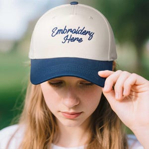 May include: A cream-colored baseball cap with a navy blue brim. The cap features the embroidered text "Embroidery Here" in a cursive font. The cap is being held by a person.
