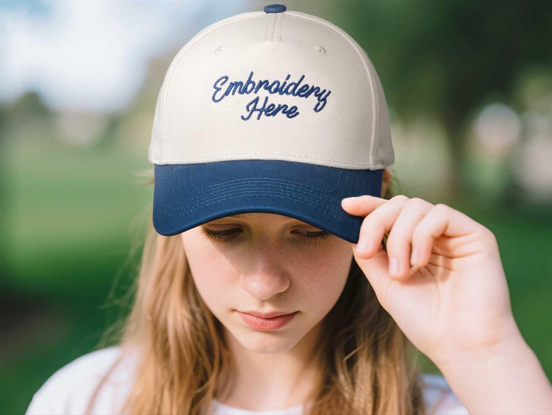 May include: A cream-colored baseball cap with a navy blue brim. The cap features the embroidered text "Embroidery Here" in a cursive font. The cap is being held by a person.