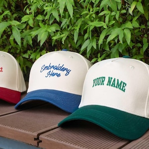 Puede incluir: Una fila de gorras de béisbol personalizables con coronas blancas y viseras de colores. Las gorras muestran texto bordado en rojo, azul y verde. El fondo presenta follaje verde.
