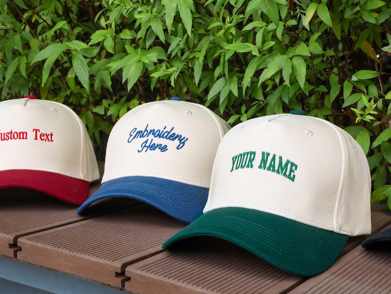 May include: A row of customizable baseball caps with white crowns and colored brims. The caps display embroidered text in red, blue, and green. The background features green foliage.