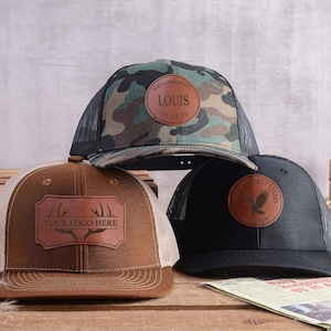 May include: Three trucker hats with leather patches. One hat is camouflage with a round patch that says "Groomman" and the name "LOUIS" and the date "07.08.20". The second hat is brown with a rectangular patch that says "YOUR LOGO HERE". The third hat is black with a round patch that says "LAST STAND".