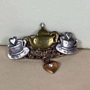 May include: A metal brooch featuring a central brass-toned teapot flanked by two silver-toned teacups on saucers. Each cup contains a small heart, and a dangling heart charm with a clear stone hangs below. The base has an intricate filigree design.