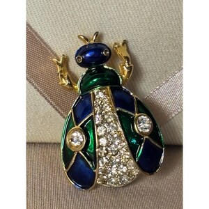 May include: A jeweled insect brooch with a blue head, green and blue body, and gold-colored accents. The brooch features a central row of clear crystals and two round crystal accents.