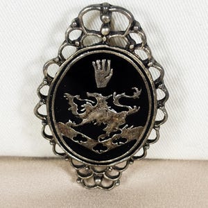 May include: An oval-shaped brooch featuring a silver-toned metal filigree frame surrounding a black center inlay. The center displays a raised silver-toned relief of a mythical dragon-like creature positioned above a chevron with three clover shapes.