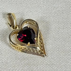 May include: A gold heart-shaped pendant featuring a deep red, faceted gemstone at its center. The outer frame of the pendant is crafted with an intricate, openwork filigree design, and it includes a gold bail at the top for attachment to a chain.