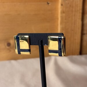 May include: A pair of rectangular earrings featuring a polished gold-tone metal finish with a smooth, dark navy blue enamel inlay forming an L-shaped geometric pattern on the surface of each piece, mounted on a black display stand.