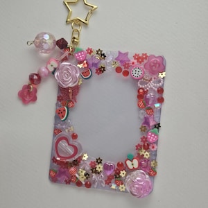 May include: A clear, rectangular photo holder decorated with pink, red, and gold embellishments. The frame is adorned with flowers, hearts, strawberries, and a star-shaped keychain. A beaded charm hangs from the top.
