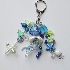 Könnte beinhalten: Ein Schlüsselanhänger mit Perlen in einem Quallen-Design, mit blauen, weißen und grünen Perlen. Der Schlüsselanhänger hat einen silberfarbenen Verschluss und Charms, darunter ein Seestern und ein Kreuz.