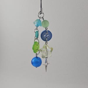 May include: A beaded charm with a variety of colorful beads and a star pendant. The charm features blue, green, and clear beads, along with a blue button and a silver-colored star. The charm is attached to a black cord.