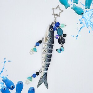 May include: A segmented fish keychain featuring a silver star-shaped ring, adorned with blue and black beads, a small butterfly charm, and additional star charms. The fish has a grey and white spotted pattern, surrounded by small blue fish-shaped beads.
