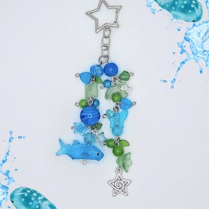 May include: A decorative keychain featuring a silver star-shaped clasp, dangling blue and green beads, small silver star charms, a blue butterfly-shaped bead, and a light blue whale shark-shaped charm, all connected by silver metal jump rings.