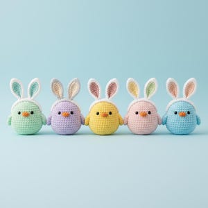 May include: Five colorful crochet chicks wearing bunny ears. The chicks are in shades of green, purple, yellow, pink, and blue. Each chick has a small orange beak and is set against a light blue background.