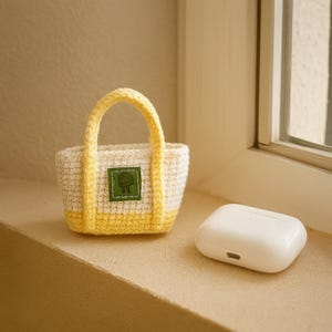 May include: A small, hand-crocheted tote bag in cream and yellow, with a green patch featuring a tree design. Next to it is a white, rectangular case, likely for earbuds. The bag has a yellow handle.