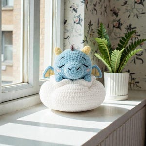 Może przedstawiać: Ręcznie robiony, szydełkowy niebieski smok z żółtymi rogami i skrzydłami spoczywa na białej poduszce w kształcie chmury. Zabawka ma zamknięte oczy, co sugeruje sen. Smok umieszczony jest na parapecie.