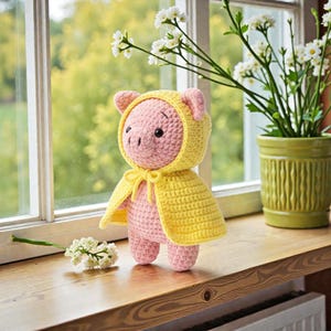 Puede incluir: Una muñeca de cerdito hecha a mano con ganchillo, principalmente rosa, con una capa con capucha amarilla atada con un lazo amarillo. El cerdito tiene ojos y orejas negros pequeños. La muñeca está de pie sobre una superficie de madera cerca de una ventana con un jarrón de flores blancas.