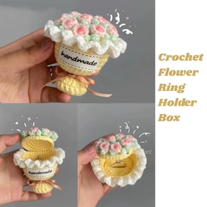 Puede incluir: Caja de ganchillo hecha a mano para guardar anillos con forma de cupcake, con un ramo de flores rosas y blancas en la parte superior. La caja es amarilla con un borde blanco con volantes y una cinta. El texto "handmade" está en una pequeña etiqueta.