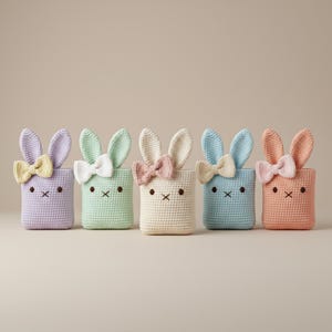 May include: Five pastel-colored crochet bunny baskets. Each basket has a different color, including lavender, mint green, cream, light blue, and peach. Each bunny basket has a bow and embroidered facial features.