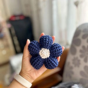Puede incluir: Una flor de ganchillo azul marino hecha a mano con un centro blanco. La flor tiene cinco pétalos y se sostiene en una mano. El trabajo de ganchillo parece estar tejido apretadamente, dando a la flor una apariencia texturizada. Es un artículo decorativo.