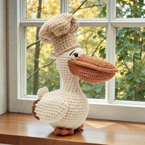 Puede incluir: Un pelícano tejido a ganchillo con un gorro de chef. El pelícano es principalmente blanquecino con pico, patas y detalles marrones. Está sobre una superficie de madera frente a una ventana con un fondo exterior borroso. Un artículo decorativo hecho a mano.