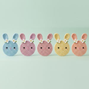 May include: Five crocheted bunny faces in pastel colors: blue, pink, light pink, yellow, and peach. Each bunny has black eyes, a stitched nose, and a small flower crown. The bunnies are arranged in a row against a light green background.
