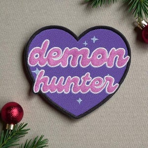May include: A purple heart-shaped patch with the words "demon hunter" in pink, outlined in white. The patch has a black border and small star accents. The background is a textured, light-colored surface.