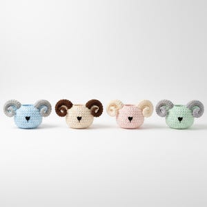 May include: Four crocheted ram-shaped containers in pastel colors: light blue, cream, pink, and mint green. Each has a heart-shaped nose and curled horns. These decorative containers are ideal for small items.