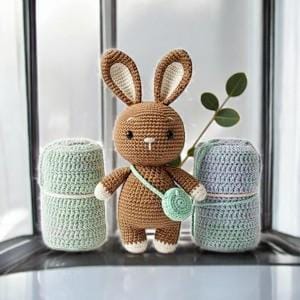 May include: A handmade crochet bunny with a small green purse, standing between two spools of yarn. The bunny is brown with cream-colored accents on its ears, feet, and hands. The yarn spools are light green and blue-gray. The scene is set near a window.