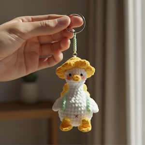 Puede incluir: Llavero de ganchillo hecho a mano con un ganso blanco que lleva un sombrero amarillo y una pequeña mochila verde. El ganso tiene patas amarillas y una pequeña cruz roja cosida en la barriga. El llavero está sujeto a un aro de metal plateado.