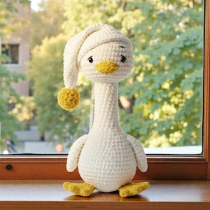 May include: A handmade, crocheted goose doll in cream and yellow. The goose wears a cream-colored nightcap with a yellow pom-pom. It has a yellow beak and feet, and black eyes. The doll is sitting on a wooden surface in front of a window.