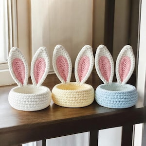 Può includere: Tre cestini a forma di coniglietto all'uncinetto in bianco, giallo e blu. Ogni cestino ha orecchie di coniglio dritte con l'interno rosa. I cestini rotondi sono su una superficie di legno scuro, perfetti per le decorazioni pasquali.