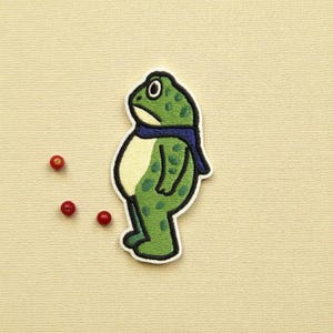 May include: A green frog patch with a blue scarf and white border. The frog has a light yellow belly and green spots. Three small red berries are scattered near the patch on a beige background.