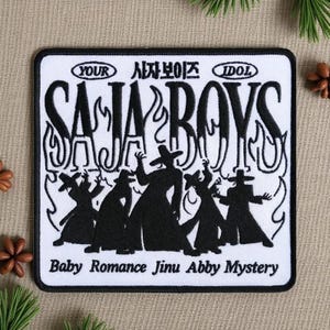 May include: A square, white patch with black embroidered text and graphics. The text reads "SAJA BOYS" in large, stylized letters, with additional text above and below. Silhouetted figures are depicted below the text.