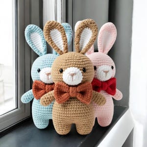 May include: Three handmade crochet bunny rabbits in blue, brown, and pink. Each bunny has a bow tie and contrasting inner ear color. The bunnies are soft and cuddly, perfect for a child's toy or nursery decor.