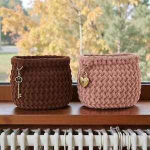 Pode incluir: Duas cestas de crochê feitas à mão em marrom e rosa, cada uma com um charme decorativo. A cesta marrom tem um charme de chave e a cesta rosa tem um charme de cadeado em forma de coração. As cestas são exibidas em uma superfície de madeira.